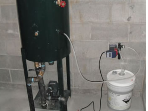 Vertical condensate water feed system with a five gallon pail of boiler chemical connected by pump and tubing.